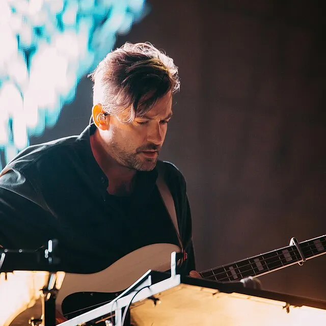 Bonobo playing bass on a dark stage, wearing an in-ear monitor, lit from behind by a pale blue backdrop