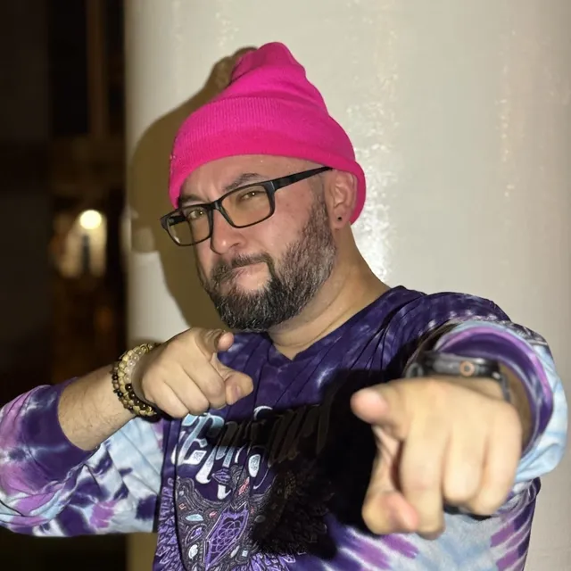 OnBrand in a bright pink beanie and black-framed glasses, wearing a purple and blue tie-dye long-sleeve shirt, pointing both index fingers straight at the camera